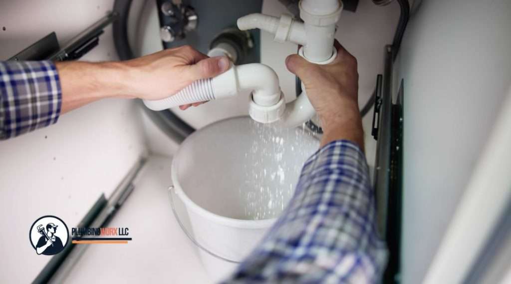 A plumber wearing a blue plaid shirt tightening a white PVC pipe under a sink with water leaking into a white bucket below, showing a plumbing repair in progress.