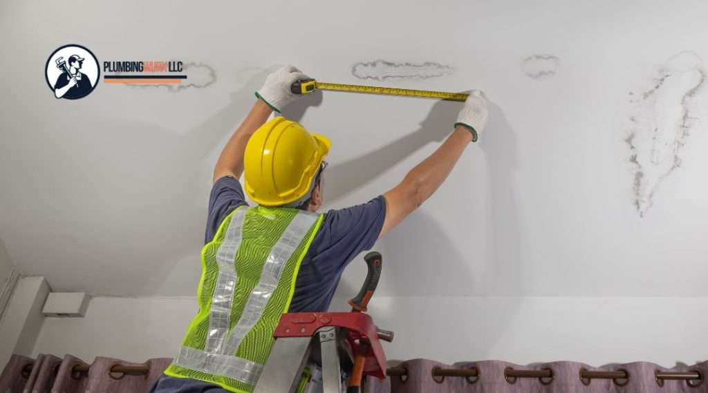 A construction worker wearing a yellow hard hat, safety glasses, and a green reflective vest measuring water damage stains on a white ceiling using a tape measure.
