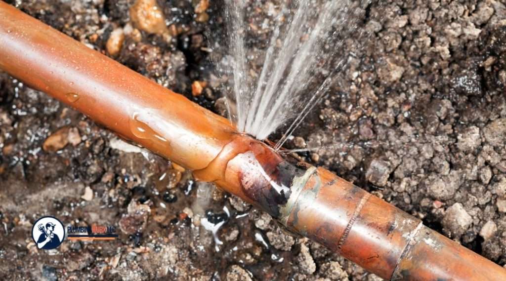Close-up of a burst copper pipe with water spraying out forcefully, surrounded by wet soil. The PlumbingWurx LLC logo is shown in the corner.