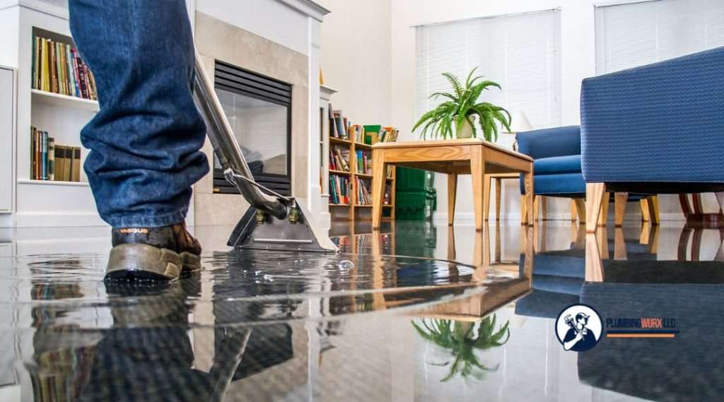 A worker uses a water extraction machine to clean up standing water on the floor in a living room, with bookshelves and furniture visible in the background.