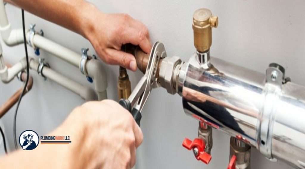 A close-up of a hand using a wrench to tighten a pipe fitting, with shiny plumbing components and valves visible.

