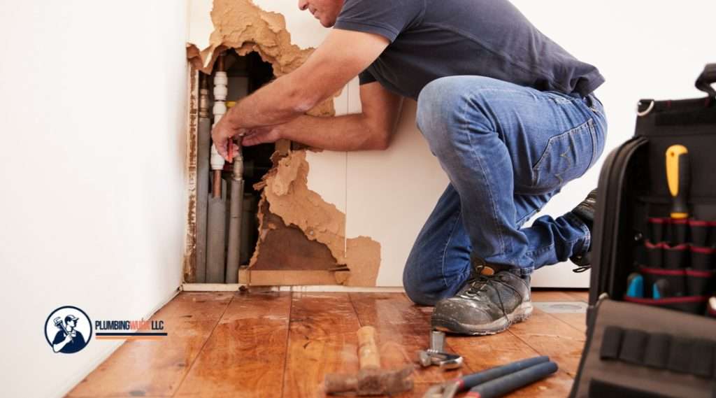 A plumber crouches down and inspects water-damaged drywall, working on pipes behind the wall, with tools nearby.